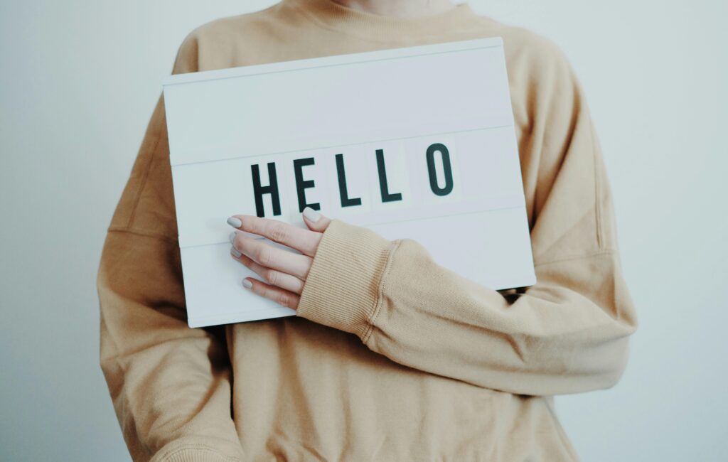 A person in beige sweater holding a 'HELLO' sign against a light background.